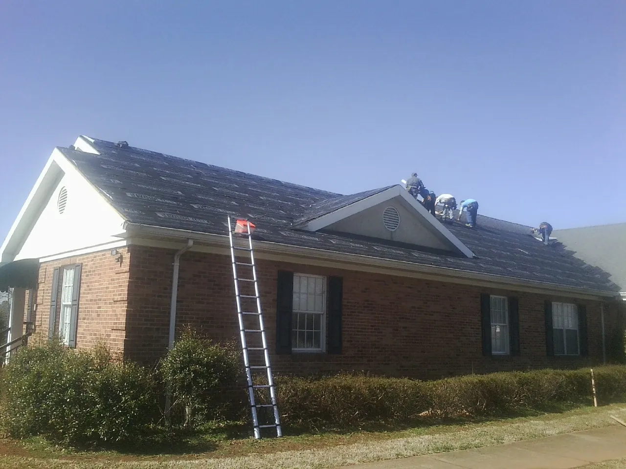 Crew tearing shingles off a brick ranch roof with a ladder set — Metal Roof Repair work by our crew in Huntersville