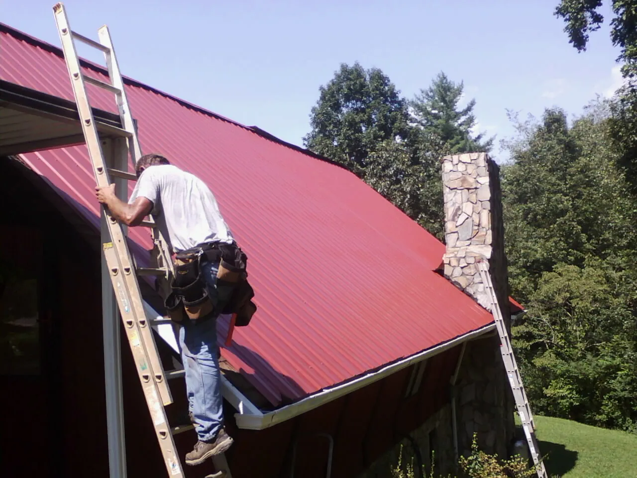 Roofer on a ladder beside a red metal roof and stone chimney — Soffit & Fascia Repair by Roofing Brothers in Huntersville
